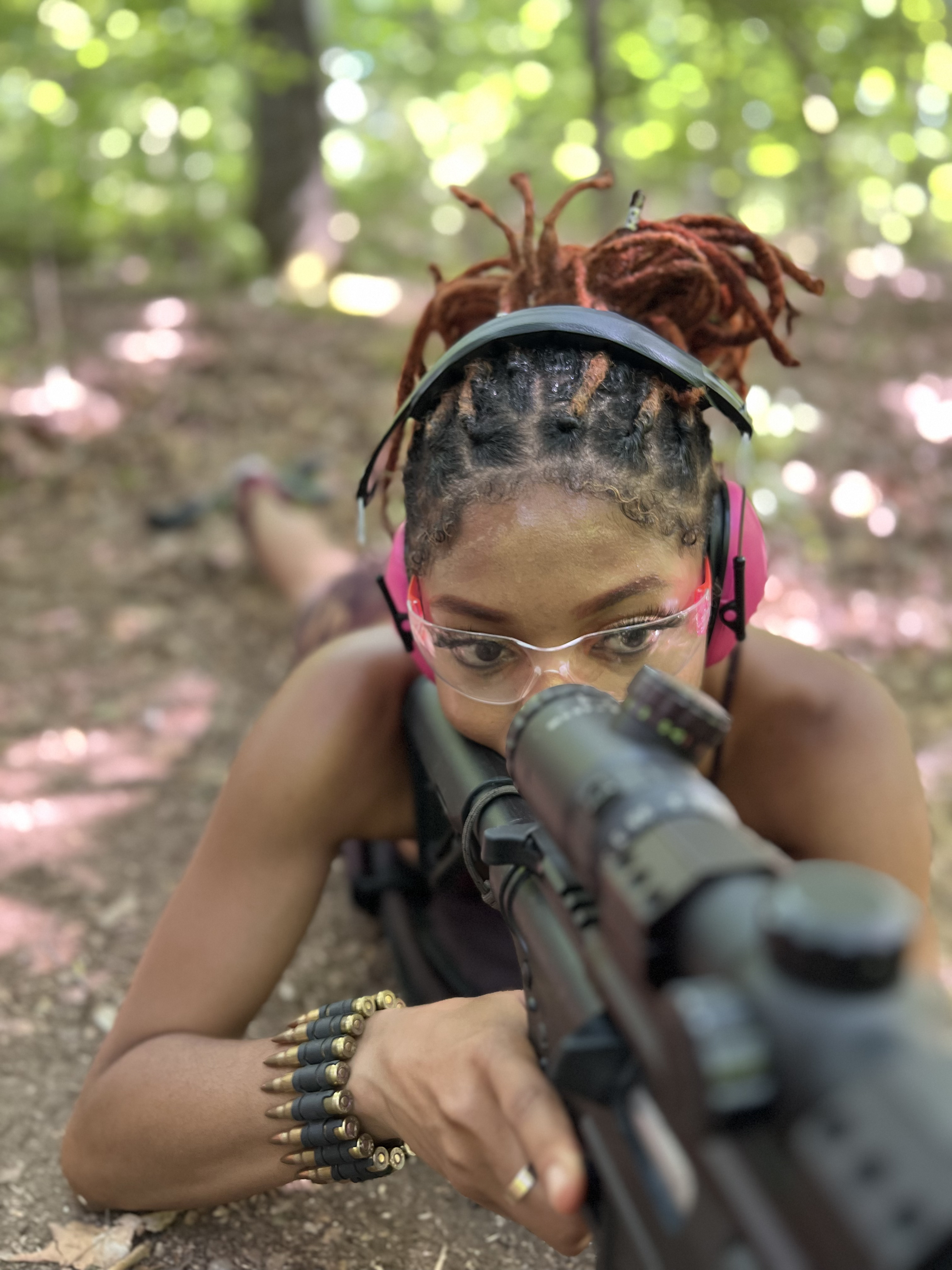 Jah The Goddess in prone position with rifle, scope, and ear protection at the gun range